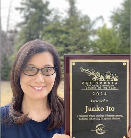 A woman with long hair and glasses holding an award plaque with green trees in the background