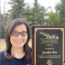 A woman with long hair and classes holding an award plaque with green trees in the background